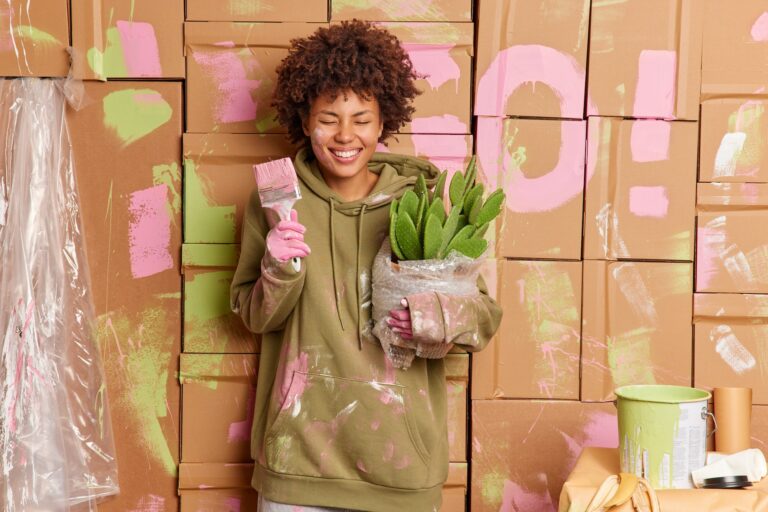 Cheerful young African American woman paints walls of living room with pink color holds cactus paint